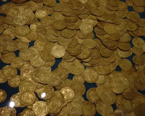 A scattered collection of ancient gold coins of varying sizes, partly embedded in soil and partly gleaming on a dark surface, with visible patina and wear
