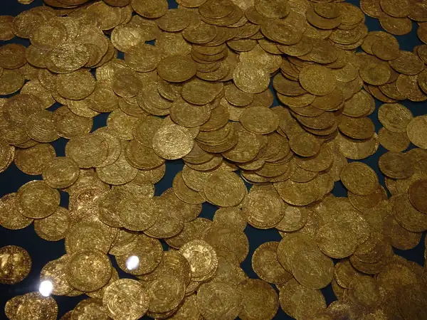 A scattered collection of ancient gold coins of varying sizes, partly embedded in soil and partly gleaming on a dark surface, with visible patina and wear