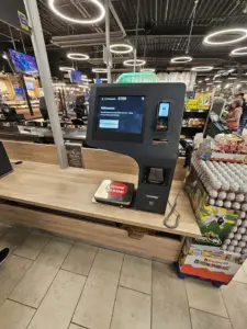 A supermarket self-checkout kiosk with touchscreen, scanner and payment terminal inside a store.
