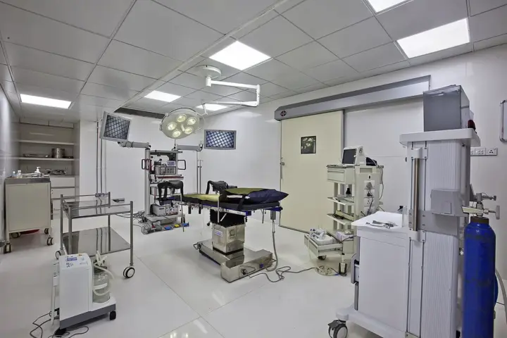 Empty hospital operating theatre with surgical table, overhead multi-head surgical lamp and anaesthesia equipment.