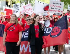 Teachers and education workers in red Australian Education Union shirts rally with megaphones and signs reading 'Fund Schools Not Sub-marines' and 'No Teachers = No Future'.