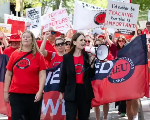 Teachers and education workers in red Australian Education Union shirts rally with megaphones and signs reading 'Fund Schools Not Sub-marines' and 'No Teachers = No Future'.