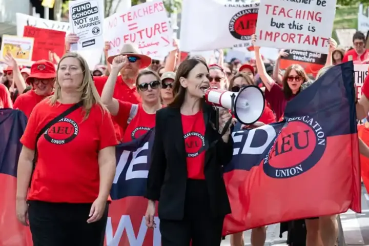 Teachers and education workers in red Australian Education Union shirts rally with megaphones and signs reading 'Fund Schools Not Sub-marines' and 'No Teachers = No Future'.