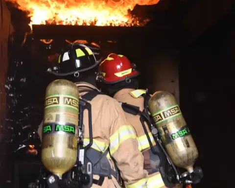 Two firefighters in full protective gear advancing into a building engulfed in flames with fire hose during emergency response