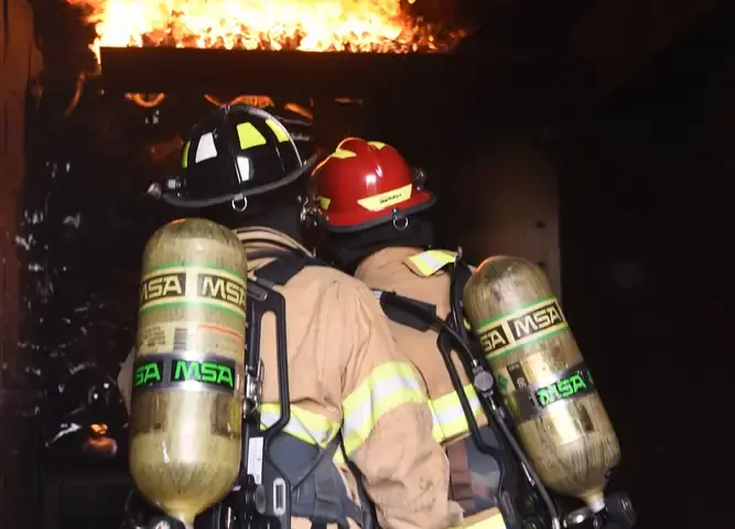 Two firefighters in full protective gear advancing into a building engulfed in flames with fire hose during emergency response