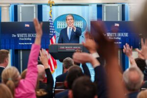 An official speaks at a White House podium during a press briefing, with presidential seal and American flags visible in the background.