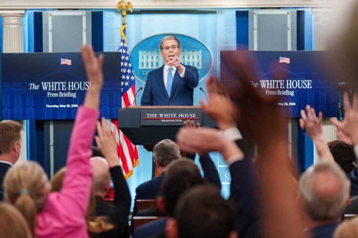An official speaks at a White House podium during a press briefing, with presidential seal and American flags visible in the background.