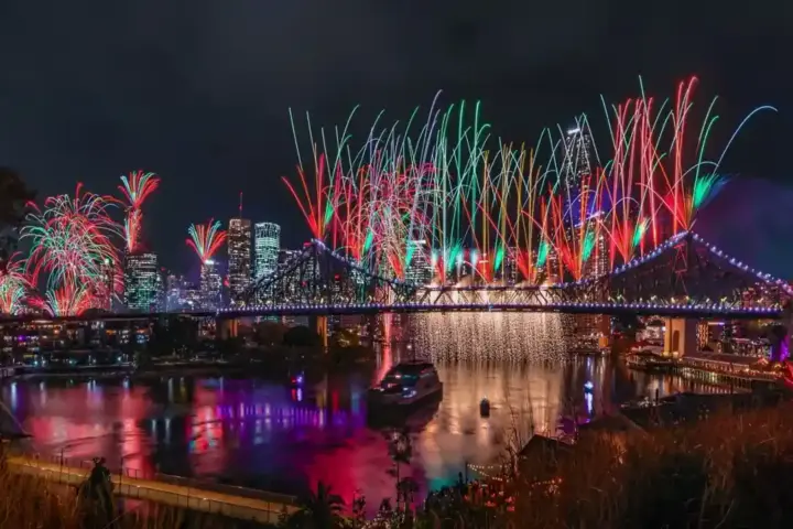 Fireworks illuminating the Brisbane River reach, bridges, and surrounding city buildings at dusk.