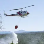 A Cal Fire helicopter performing an aerial water drop directly over dry brush in a hilly wildland terrain.