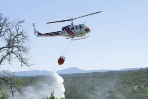 A Cal Fire helicopter performing an aerial water drop directly over dry brush in a hilly wildland terrain.