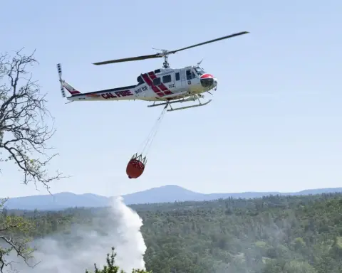 A Cal Fire helicopter performing an aerial water drop directly over dry brush in a hilly wildland terrain.