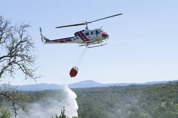 A Cal Fire helicopter performing an aerial water drop directly over dry brush in a hilly wildland terrain.