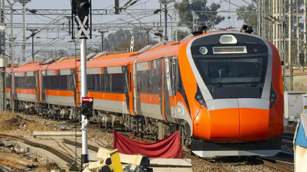 Front three-quarter angle of an aerodynamic white and blue Vande Bharat electric multiple unit traveling along overhead-electrified railway tracks.