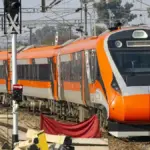 Front three-quarter angle of an aerodynamic white and blue Vande Bharat electric multiple unit traveling along overhead-electrified railway tracks.
