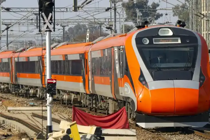 Front three-quarter angle of an aerodynamic white and blue Vande Bharat electric multiple unit traveling along overhead-electrified railway tracks.