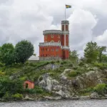 Historic castle-like structure on a rocky hill with a Swedish flag flying from the tower, cloudy sky