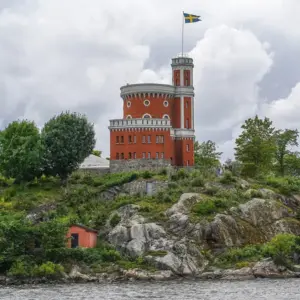 Historic castle-like structure on a rocky hill with a Swedish flag flying from the tower, cloudy sky