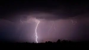 Multiple lightning bolts striking down from indigo storm clouds at night, power transmission towers silhouetted below