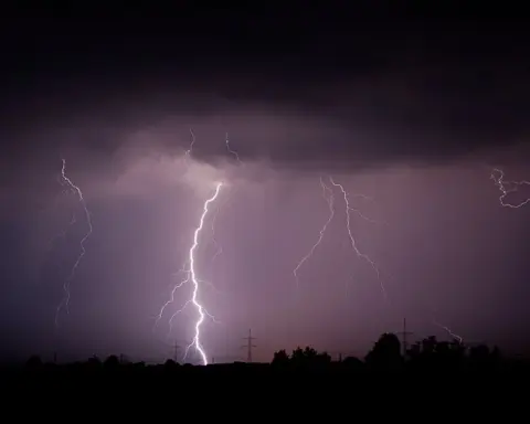 Multiple lightning bolts striking down from indigo storm clouds at night, power transmission towers silhouetted below