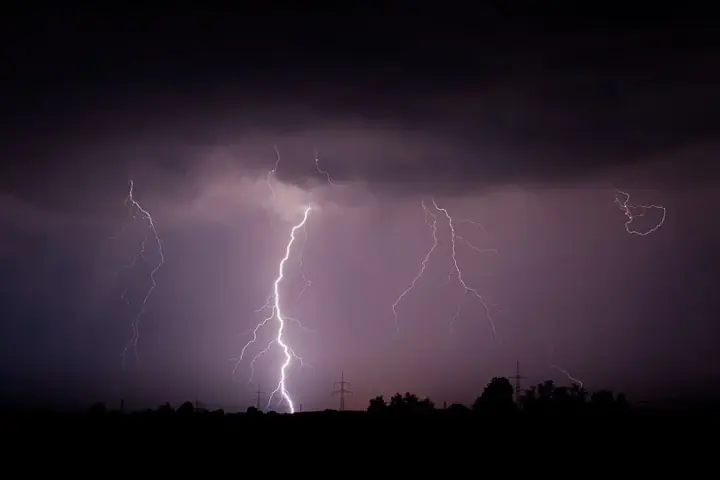 Multiple lightning bolts striking down from indigo storm clouds at night, power transmission towers silhouetted below