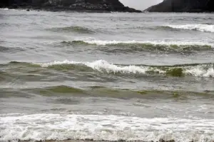 Ocean waves crashing onto a beach with white foam, dark green water, and rugged cliffs in the background under gray overcast sky