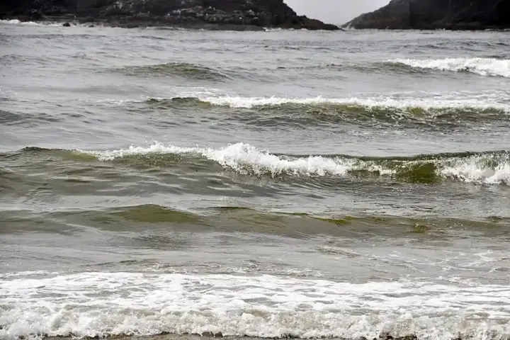Ocean waves crashing onto a beach with white foam, dark green water, and rugged cliffs in the background under gray overcast sky