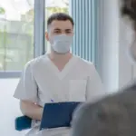 A masked healthcare professional speaks with a patient during a consultation, representing expanded Pharmacy First treatment access.