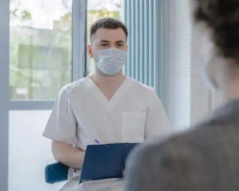 A masked healthcare professional speaks with a patient during a consultation, representing expanded Pharmacy First treatment access.