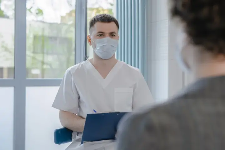 A masked healthcare professional speaks with a patient during a consultation, representing expanded Pharmacy First treatment access.