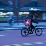 Food delivery worker riding a bicycle through New York City traffic with buildings and vehicles nearby.