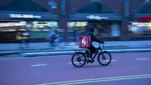 Food delivery worker riding a bicycle through New York City traffic with buildings and vehicles nearby.