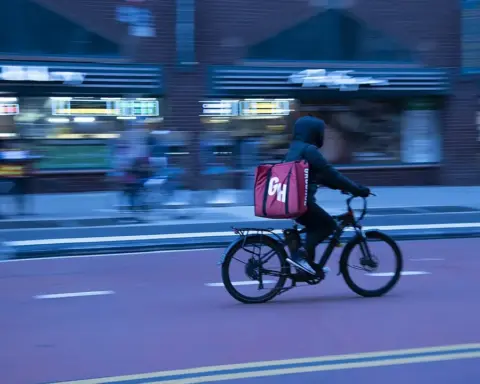 Food delivery worker riding a bicycle through New York City traffic with buildings and vehicles nearby.