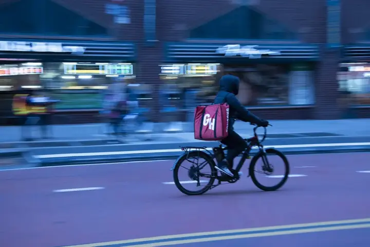 Food delivery worker riding a bicycle through New York City traffic with buildings and vehicles nearby.