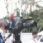 Professional broadcast video camera on a tripod with a large telephoto lens, used for documentary reporting context.