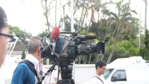 Professional broadcast video camera on a tripod with a large telephoto lens, used for documentary reporting context.