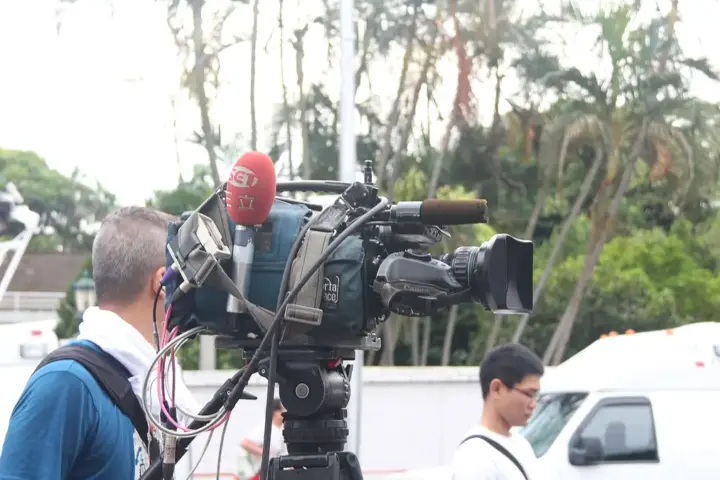 Professional broadcast video camera on a tripod with a large telephoto lens, used for documentary reporting context.