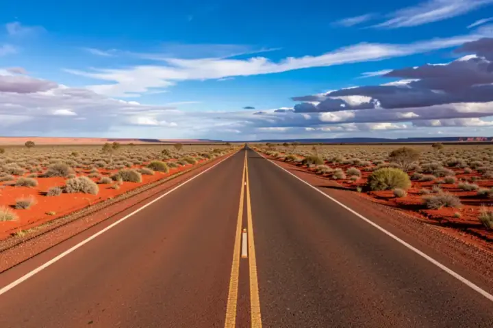 Two-lane asphalt road of the Stuart Highway cutting through arid red soil and low desert shrubs in Central Australia.