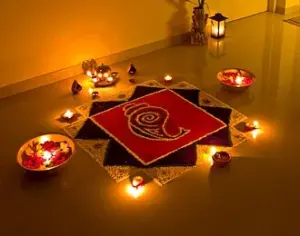 A colourful floor rangoli surrounded by lit clay diyas and brass bowls of flower petals during Diwali