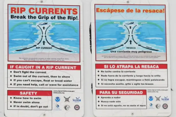 A metal warning sign posted in beach sand advising swimmers on rip current survival alongside ocean waves.