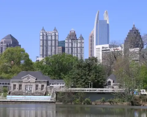 High-rise towers of the Midtown Atlanta skyline reflected across the surface of Clara Meer lake in Piedmont Park under a humid summer sky.
