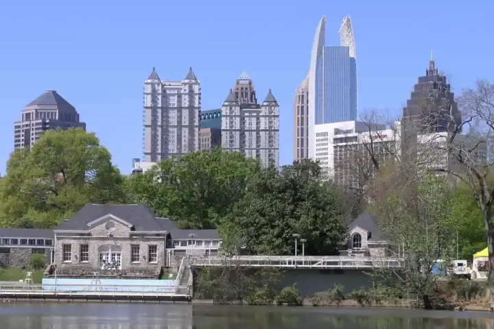 High-rise towers of the Midtown Atlanta skyline reflected across the surface of Clara Meer lake in Piedmont Park under a humid summer sky.