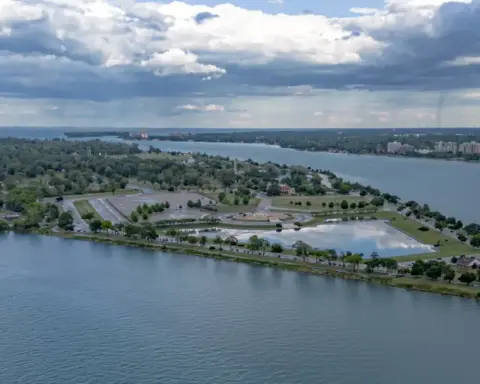 Wide view of the green grounds and Detroit River waterfront along Belle Isle Park facing toward the city skyline.