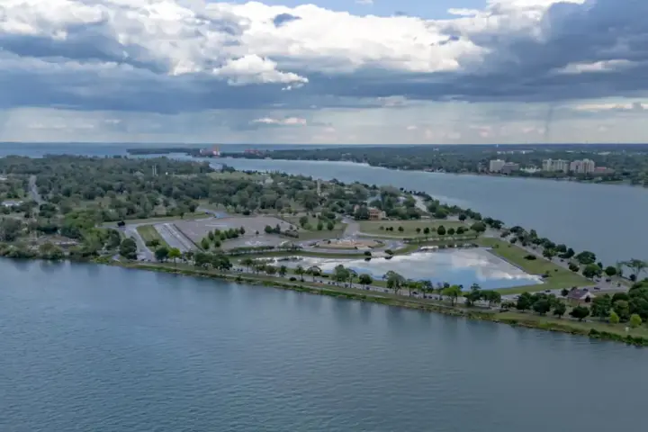 Wide view of the green grounds and Detroit River waterfront along Belle Isle Park facing toward the city skyline.