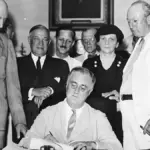 President Franklin D. Roosevelt signs the Social Security Act with officials gathered around a desk.