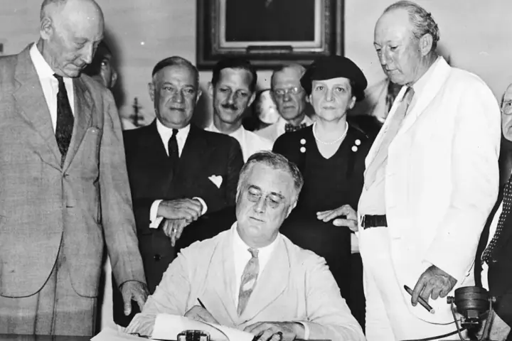 President Franklin D. Roosevelt signs the Social Security Act with officials gathered around a desk.