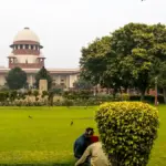 Exterior facade and central wing of the Supreme Court of India in New Delhi viewed across the institutional grounds and landscaped gardens.