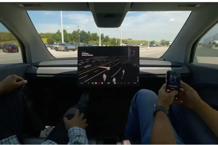 Interior view of a Tesla Cybercab showing the central screen and two passengers, with no steering wheel or foot pedals visible.