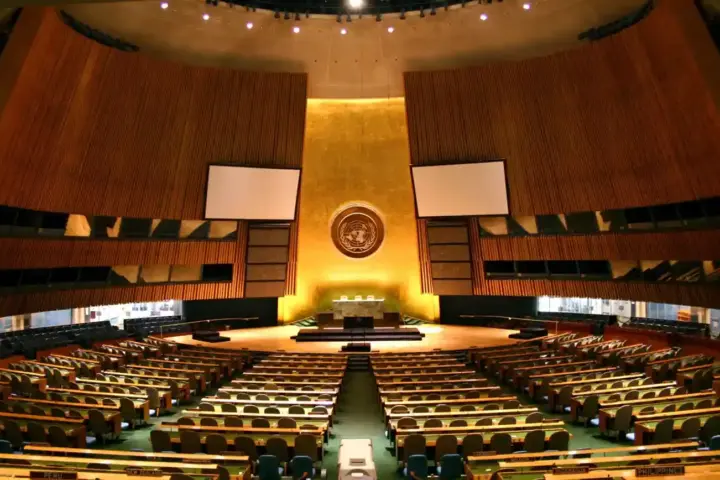 Broad interior view of the United Nations General Assembly hall showing green desks, delegation seating, and the golden podium emblem in New York City.