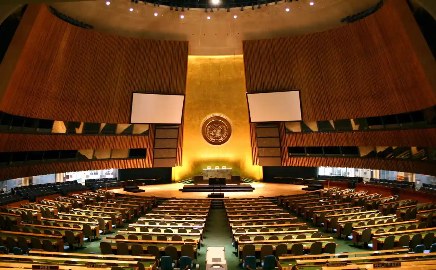 Broad interior view of the United Nations General Assembly hall showing green desks, delegation seating, and the golden podium emblem in New York City.