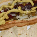 Prepared hot dog with mustard, diced onion, jalapeño and relish on a white plate.